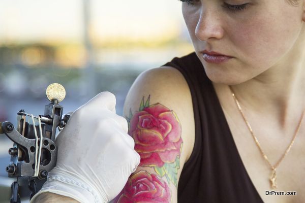 tattooer showing process of making a tattoo on young beautiful hipster woman with red curly hair arm. Tattoo design in the form of rose